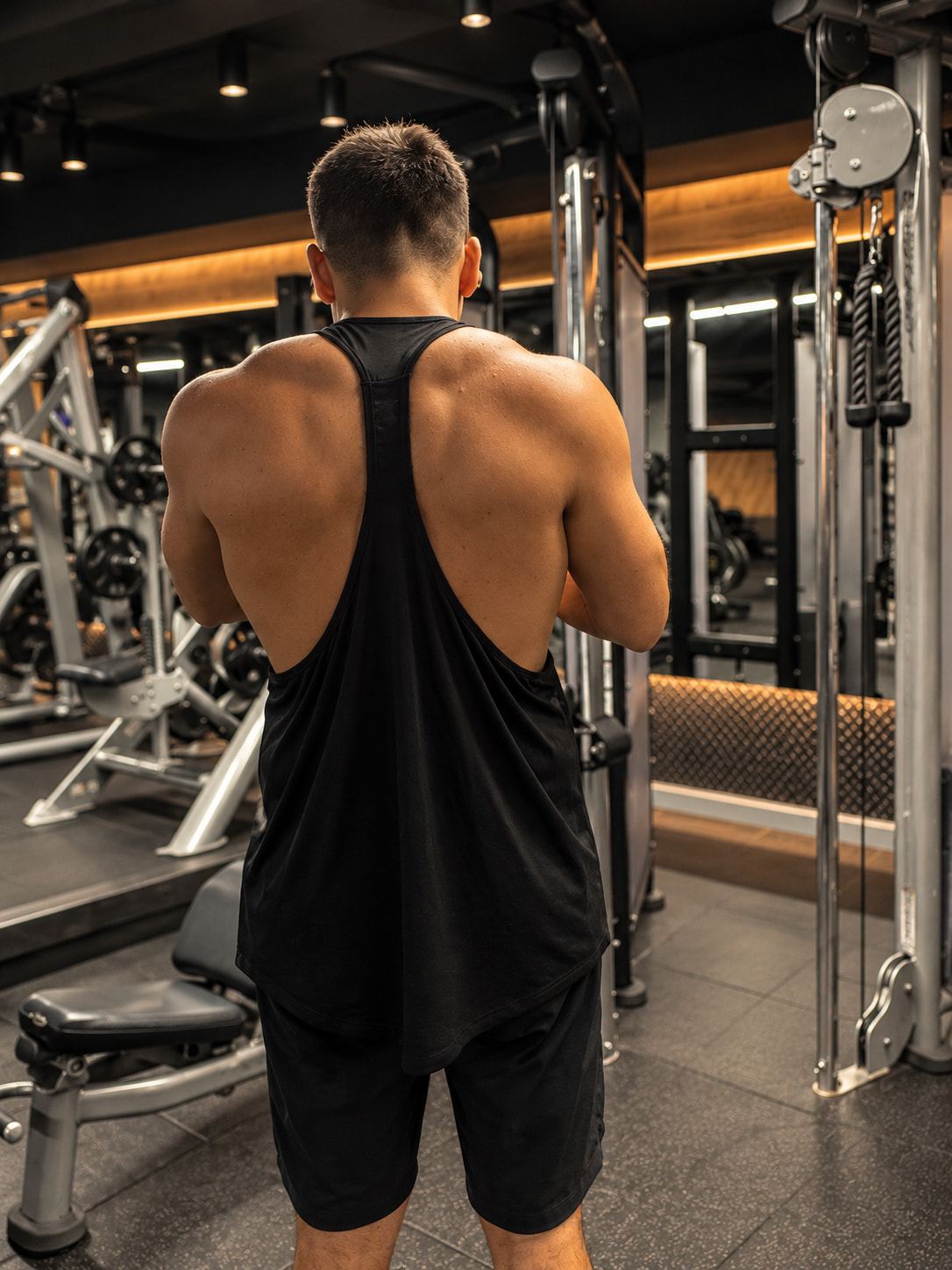 Pablo seen from behind in a black vest, showing his back and shoulders in a dim gym.
